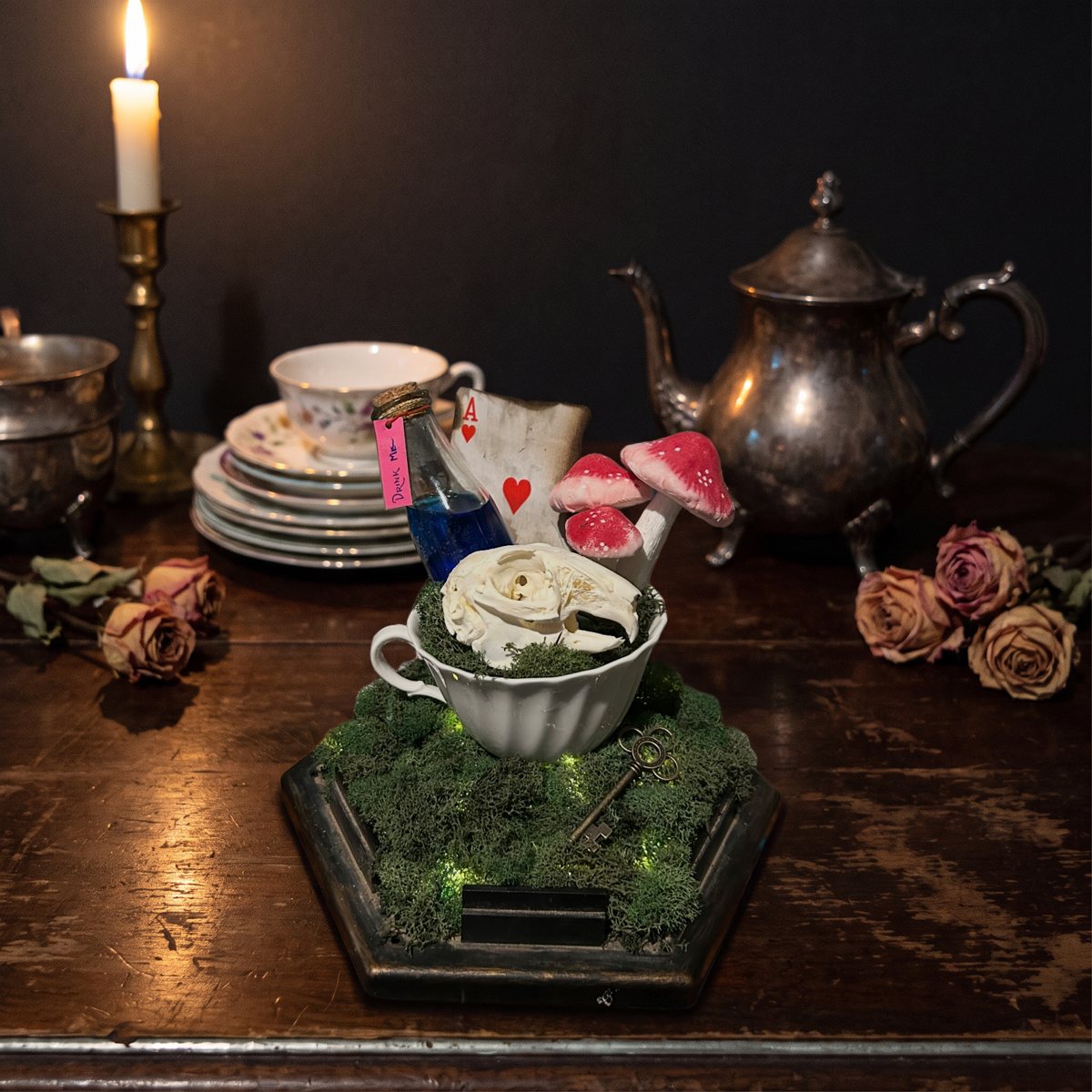The finished hero shot: the same teacup piece on a dark walnut tea table by candlelight, with a tarnished silver teapot, stacked saucers and dried roses out of focus behind it
